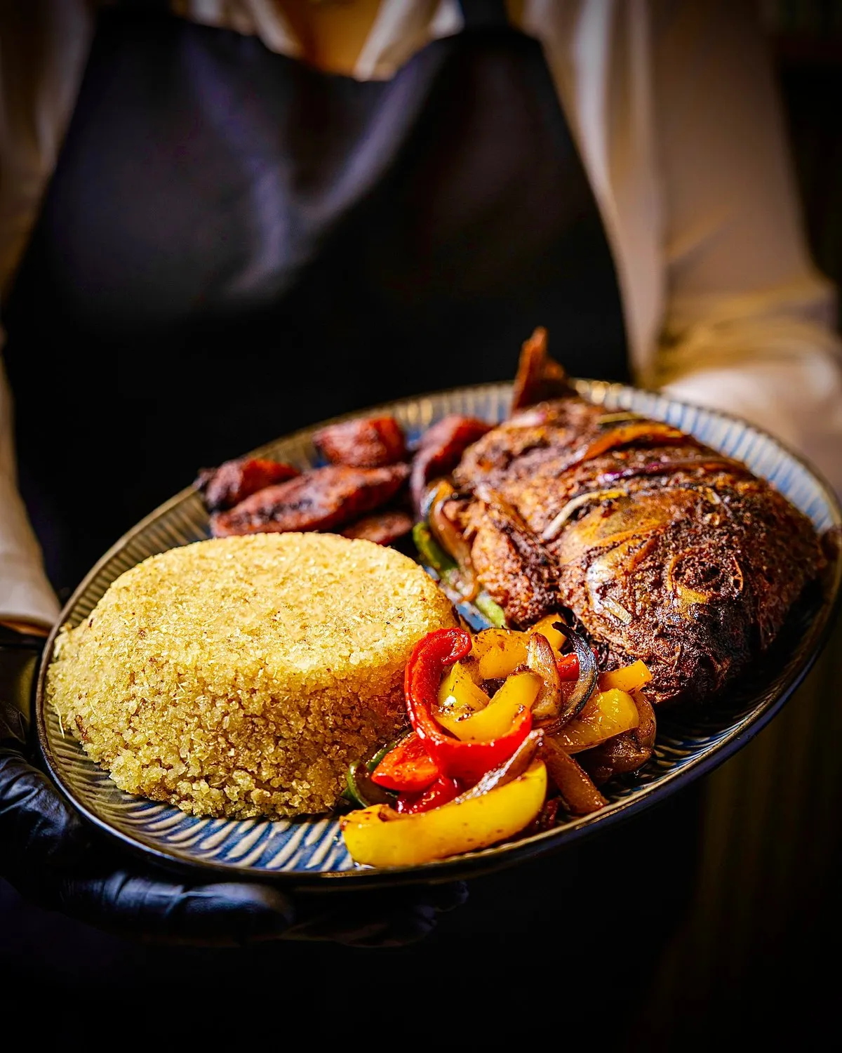 Server holding a platter of fried whole fish with couscous, peppers and plantain at Fambul.