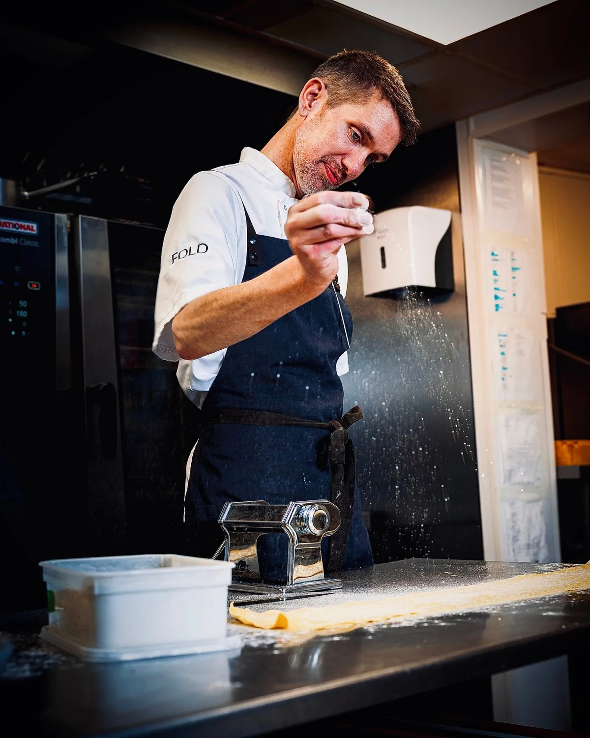 Chef dusting flour over a sheet of fresh pasta beside a pasta machine at Fold Bistro.