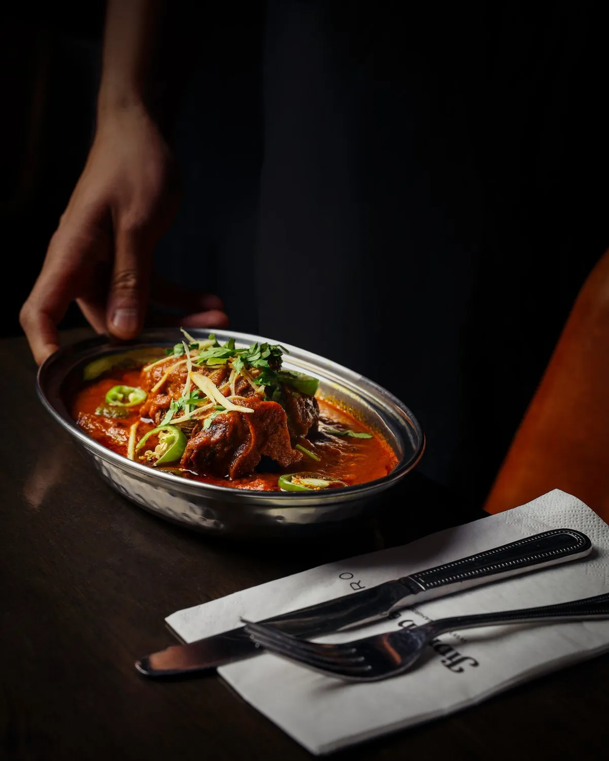Server setting down a steel karahi of lamb curry topped with ginger, coriander and green chilli at Jinnah Bistro.