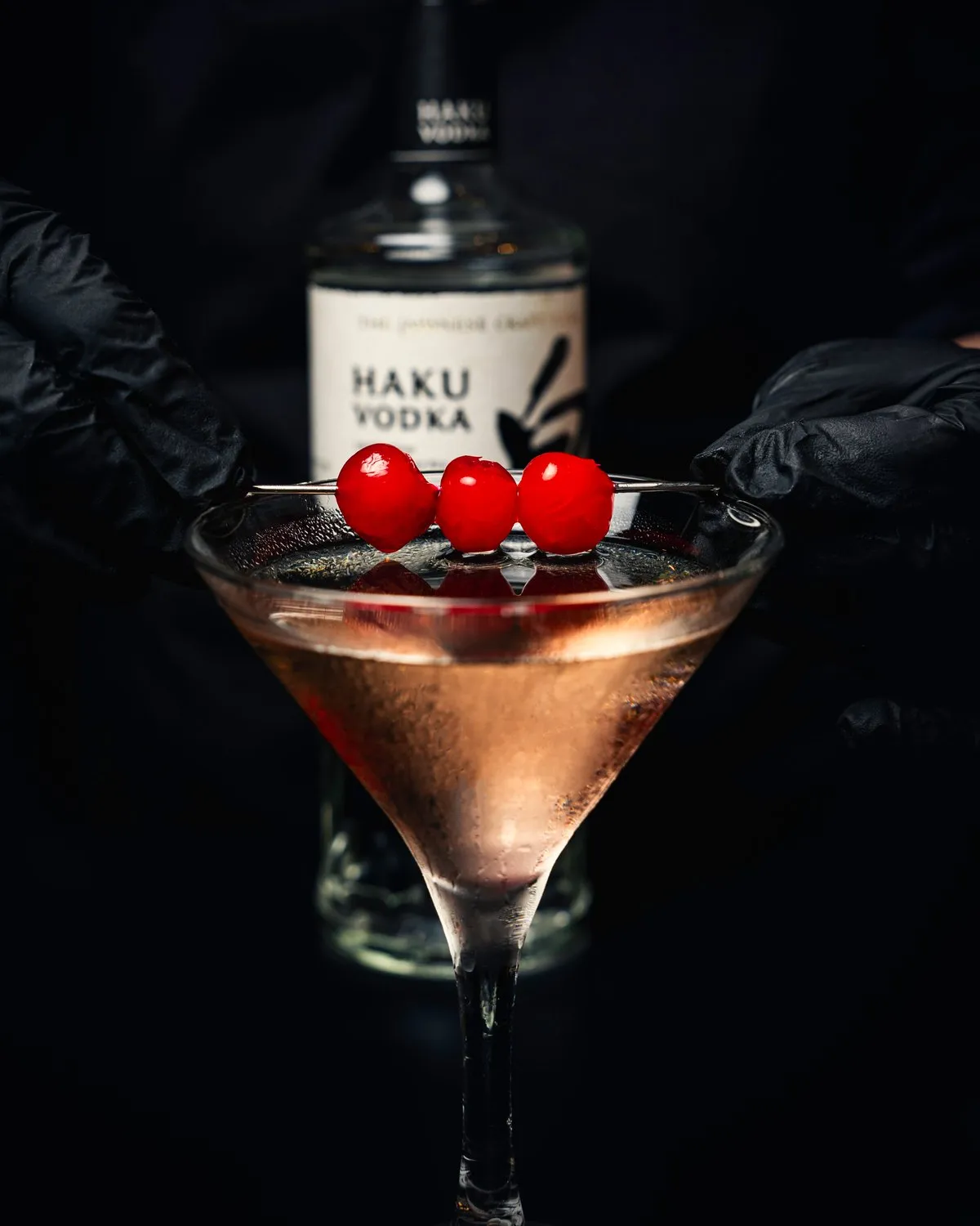 Bartender presenting a cocktail garnished with cherries beside a bottle of Haku vodka at Mina's Table.
