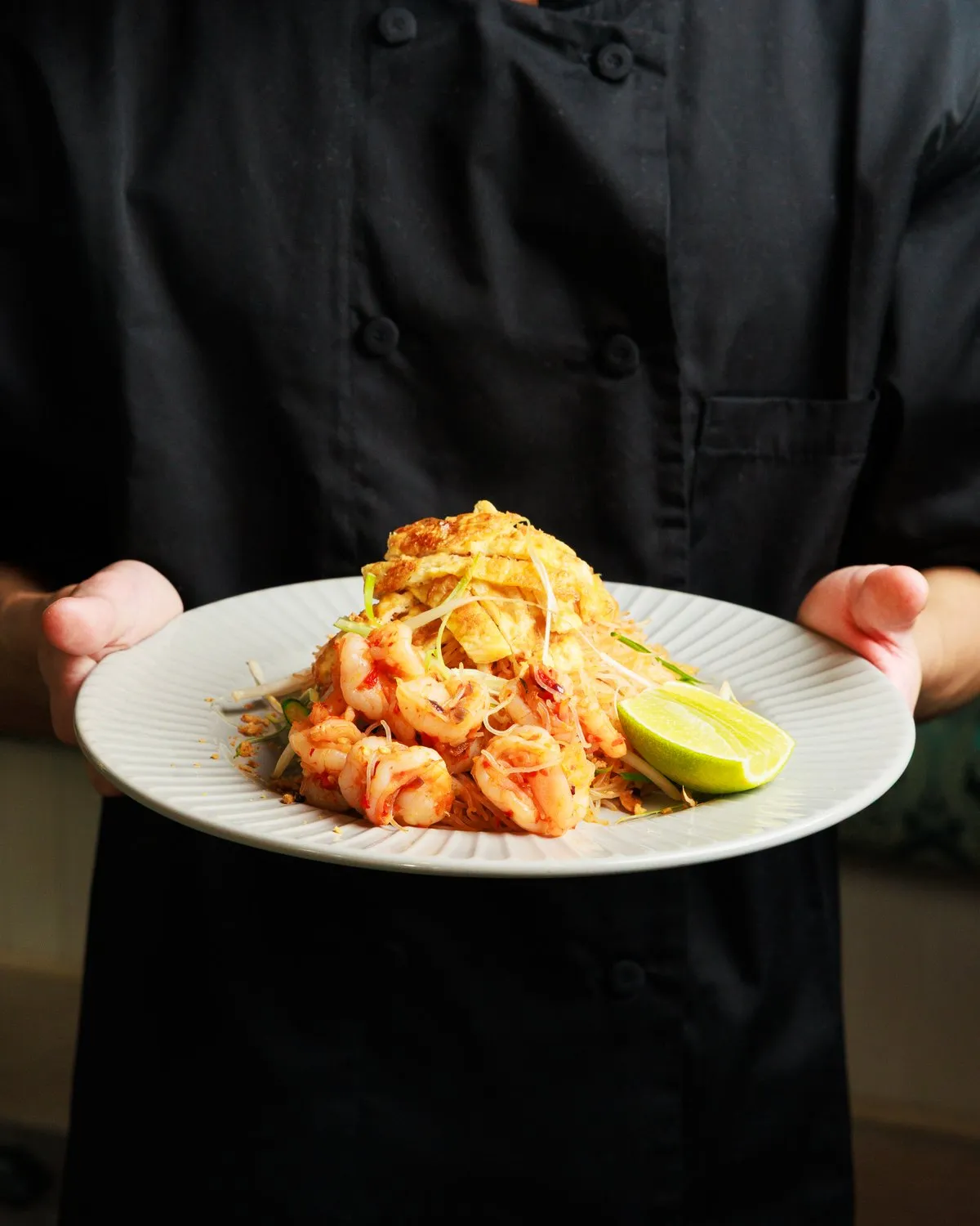 Chef holding a plate of prawn pad thai garnished with lime at Mina's Table.