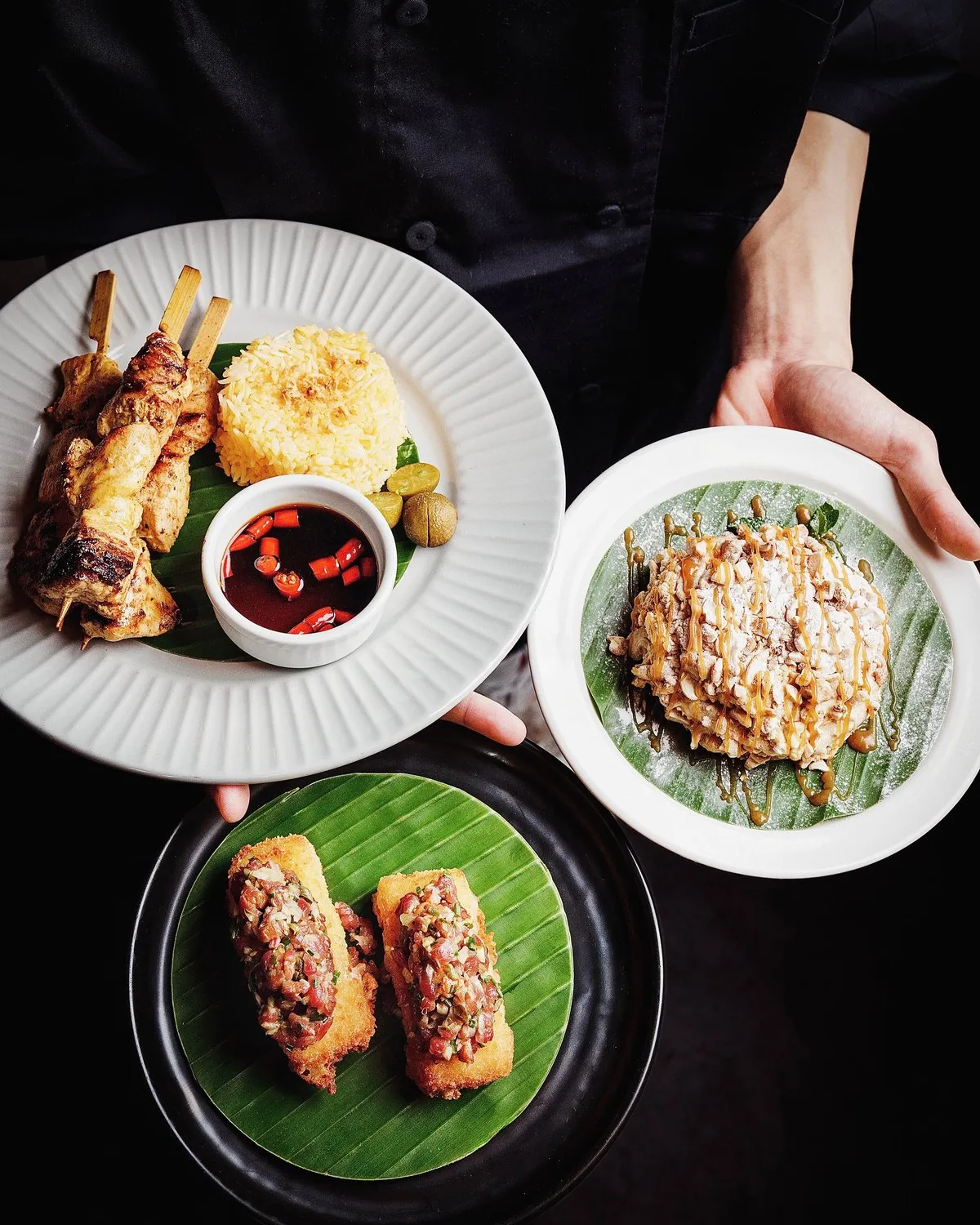 Three sharing plates of satay skewers, rice and spring rolls on banana leaves at Mina's Table.