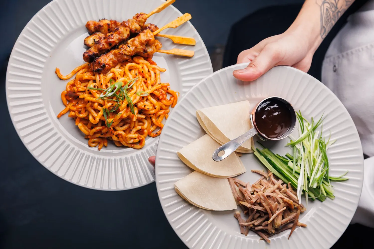 Server carrying plates of spicy noodles with chicken skewers and duck pancakes at Mina's Table.