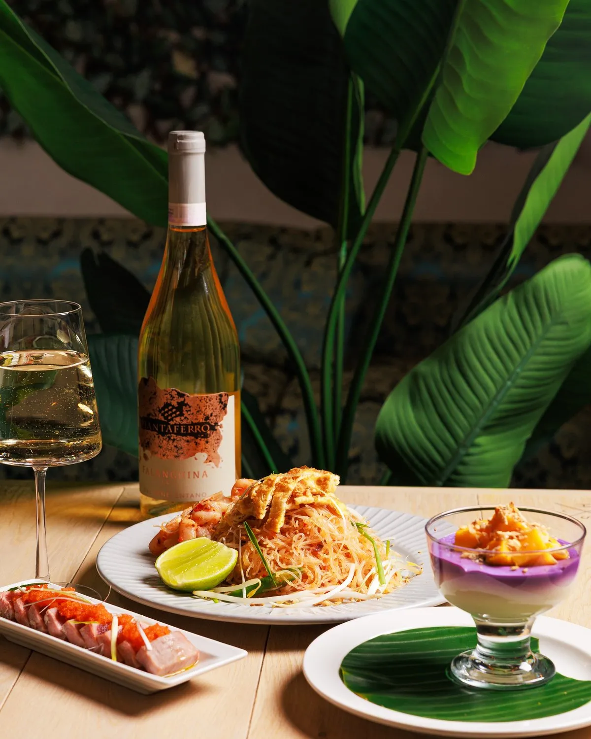 Table set with a bottle of rosé, pad thai, sashimi and a layered dessert beside tropical foliage at Mina's Table.