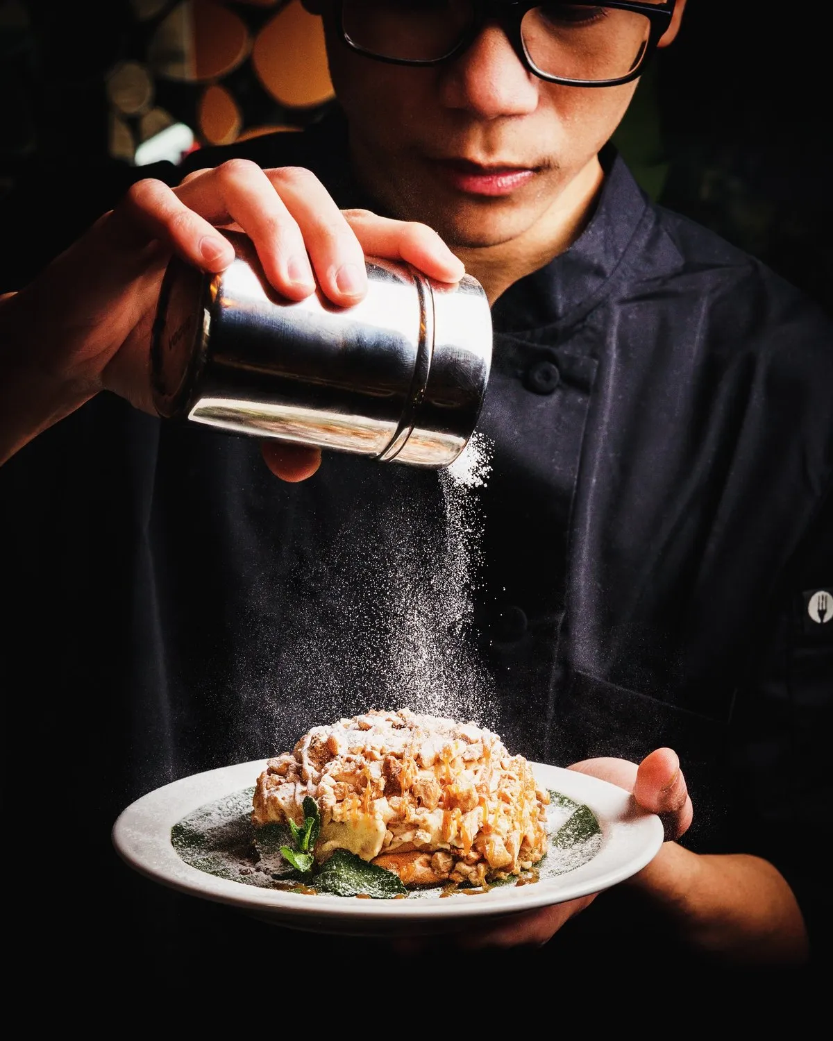 Chef dusting icing sugar from a shaker over a plated dessert at Mina's Table.