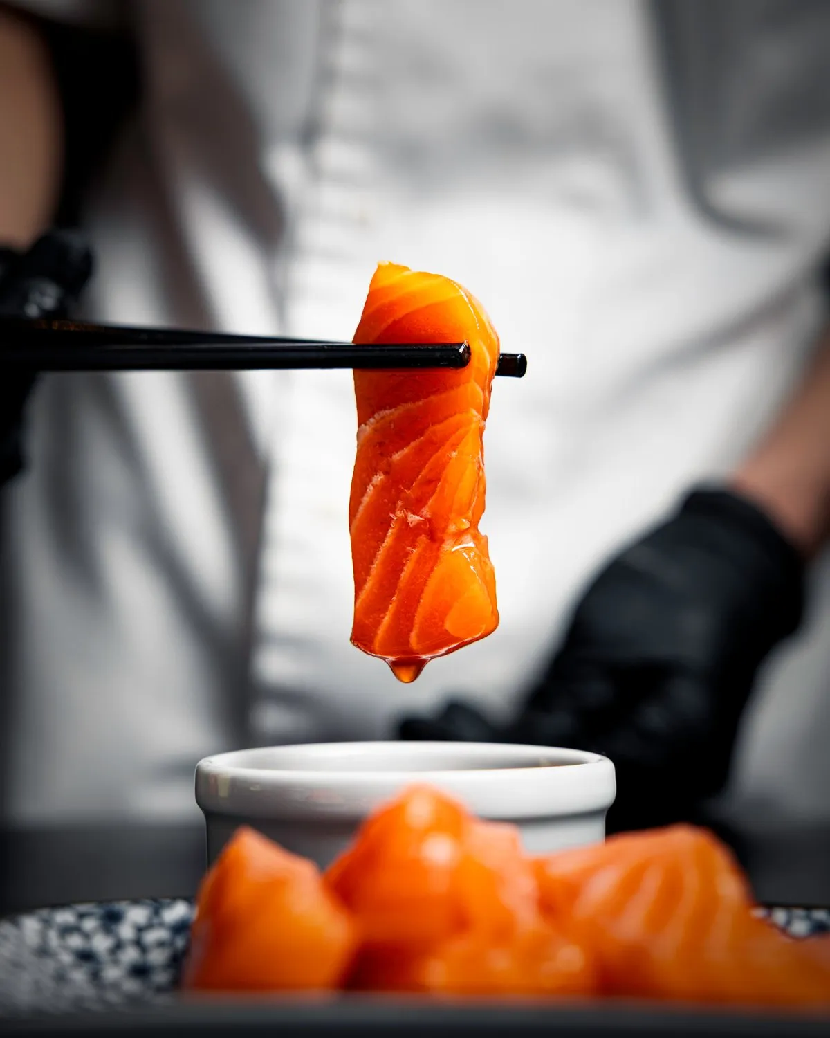 Chef lifting a slice of salmon sashimi with chopsticks above a dish of dressing at Minastable.