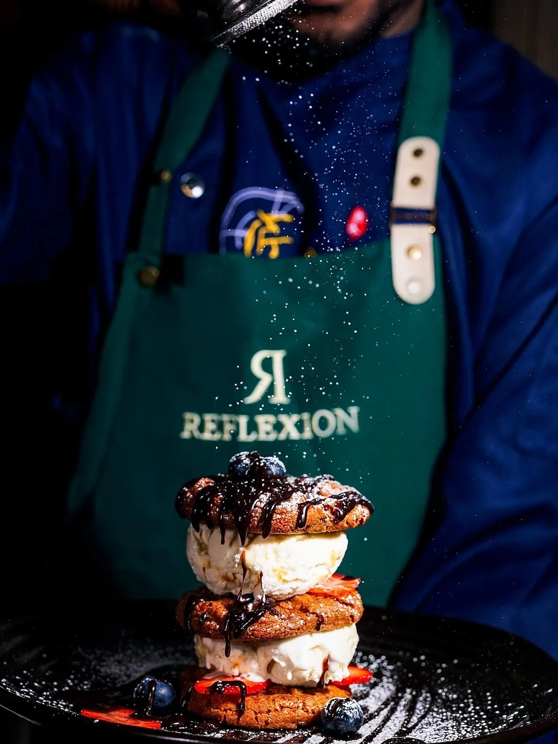 Chef dusting icing sugar over a stacked cookie-and-ice-cream dessert at Reflexion Lounge.