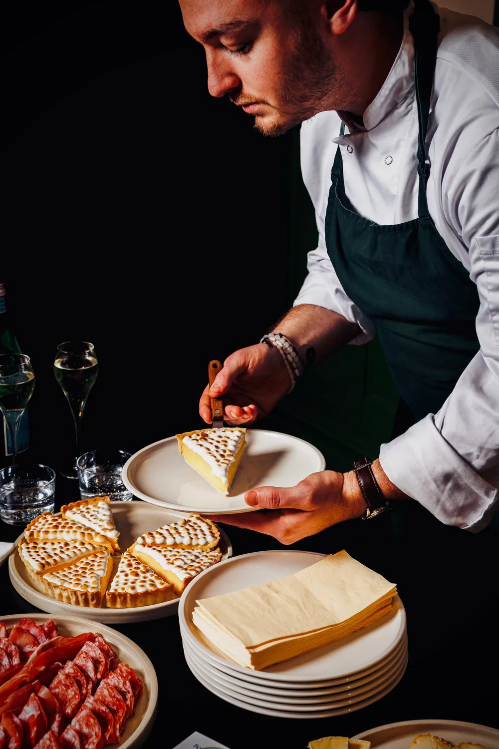 Chef plating a slice of meringue-topped lemon tart beside charcuterie and champagne at a Soothefood event.